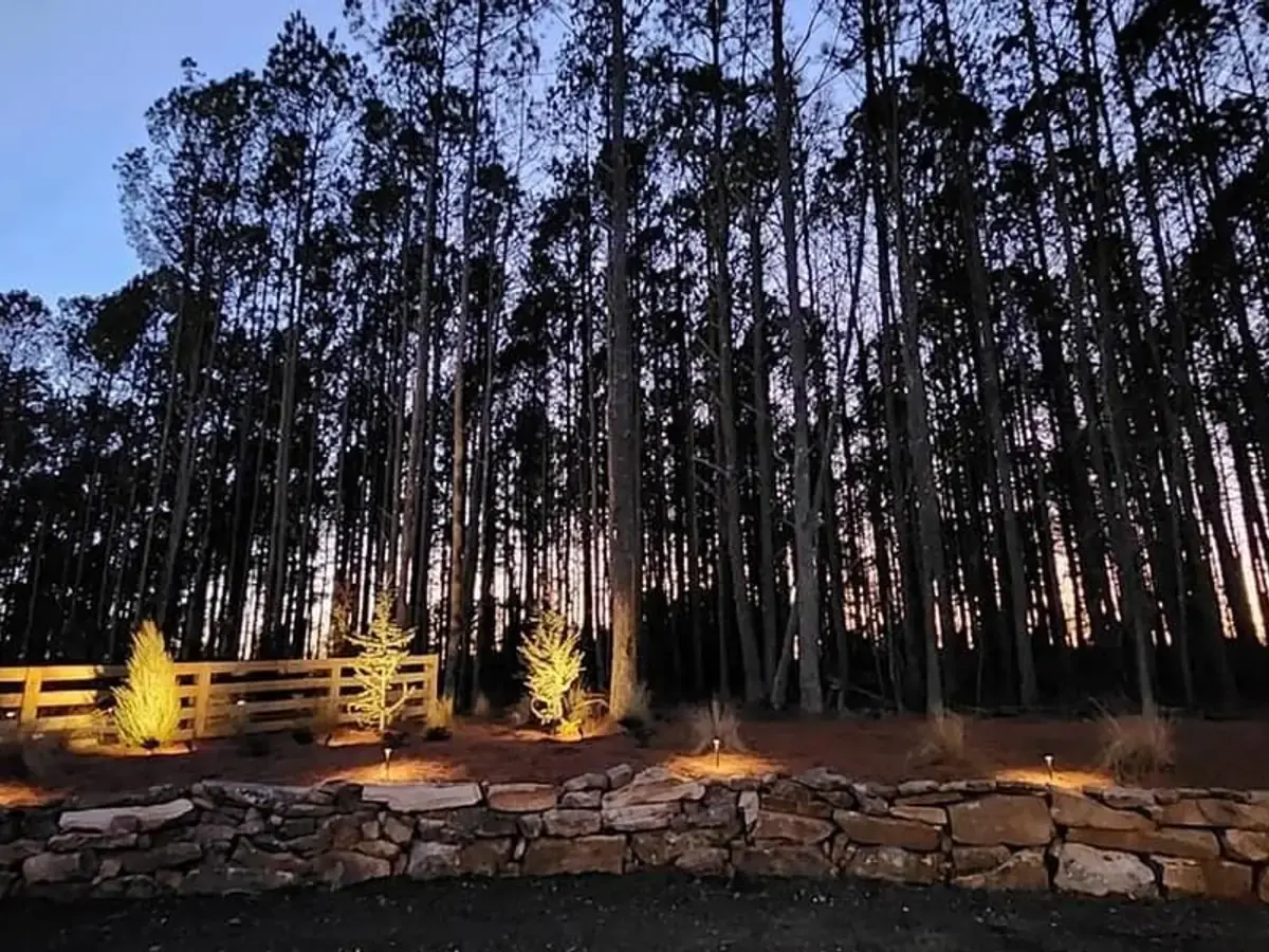 Four rail wood fence lit at dusk above a stone retaining wall, with pine trees behind it