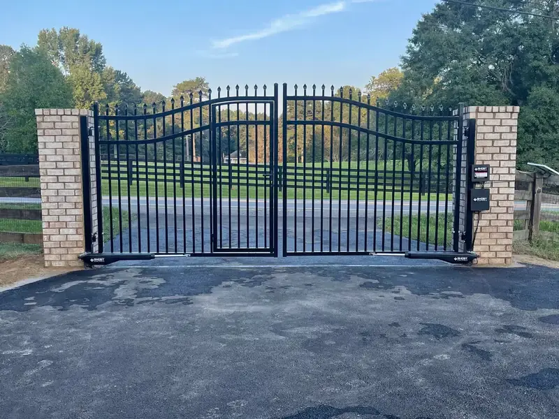 Arched black driveway gate between brick columns at the end of a paved driveway