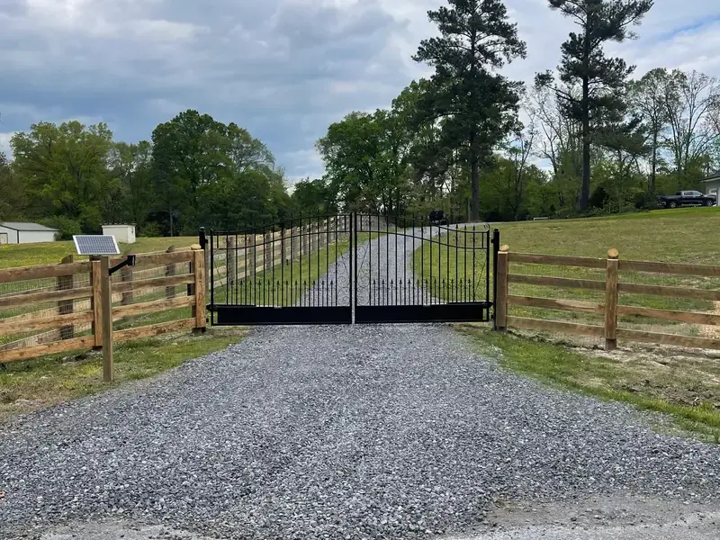 Black driveway gate set into a four rail wood ranch fence with a solar panel on the post