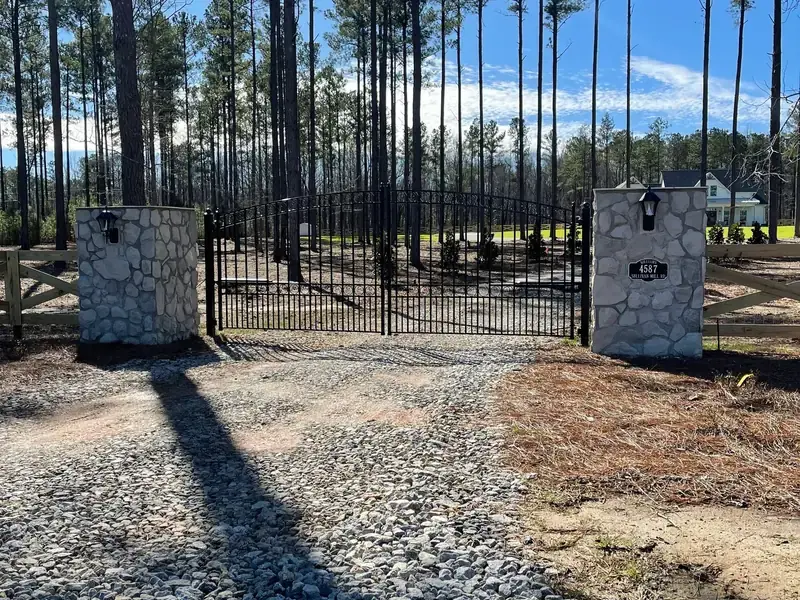 Black driveway gate hung between stacked stone columns with pine trees behind
